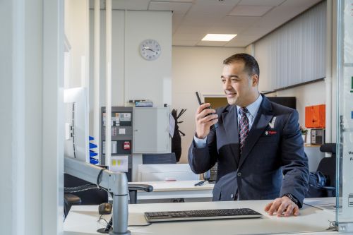 Mannelijke beveiliger in uniform praat via de portofoon met een collega beveiliger op locatie en kijkt daarnaast ook gelijk op zijn computerscherm achter zijn bureau.