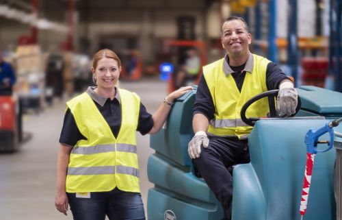 Twee schoonmaakmedewerkers van Gom Logistiek kijken lachend in de camera, 1 hiervan zit op een schrobzuigmachine, de ander staat ernaast