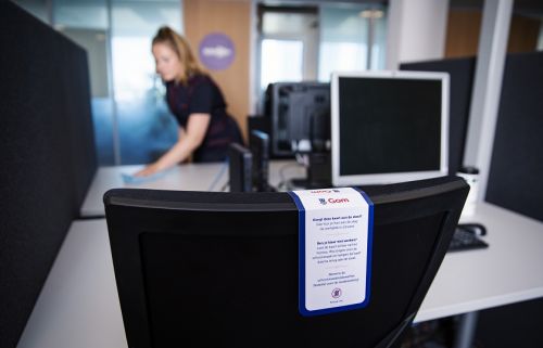 een sleeve wat aangeeft dat de flexplek schoon is, hangt over de bureaustoel, op de achtergrond maakt de schoonmaakster een ander bureau schoon