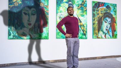 Een portretfoto van Gom Uitblinker Edmar Janga met 3 schilderijen op de achtergrond en een schaduw van een superheld met cape achter Edmar