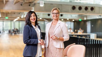 Twee vrouwen in zakelijke kleding staan geposeerd voor de camera in een lichte ruimte.