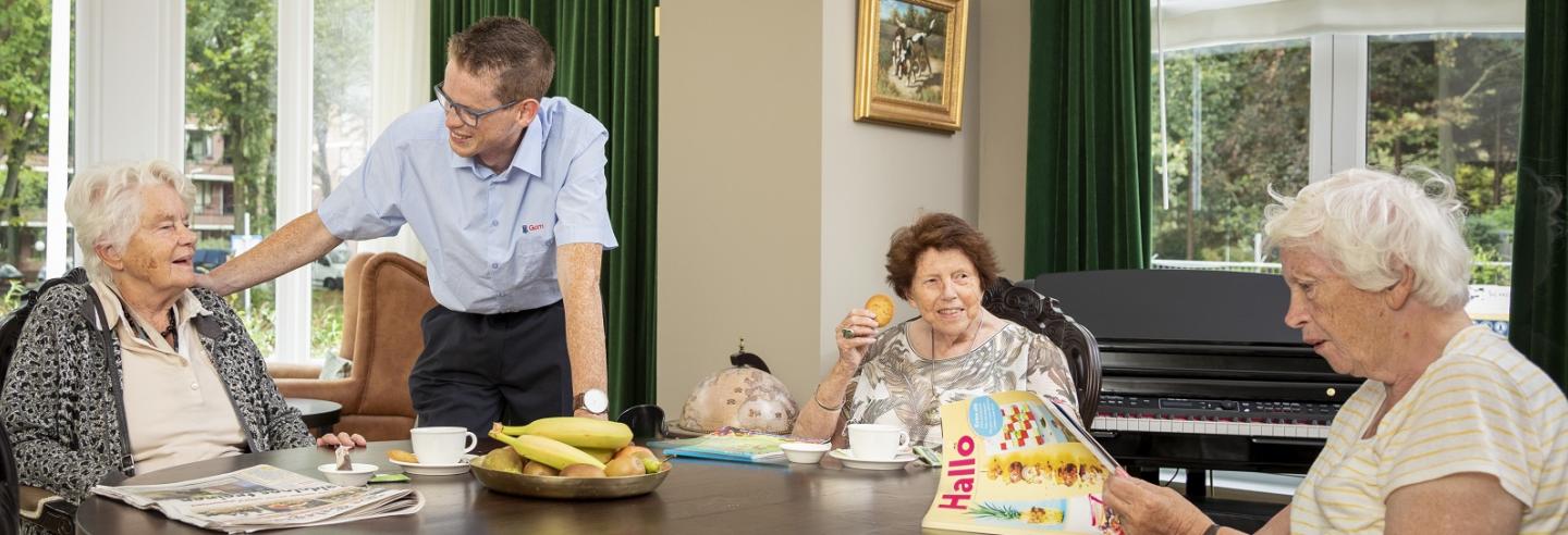 drie bejaarden vrouwen zitten aan tafel met een kopje koffie en een koekje en een schoonmaakmedewerker van Gom maakt met hen een praatje