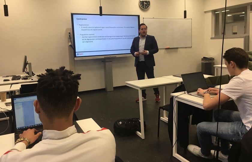 David van Boheemen staat als gastdocent voor een groot digitaal schoolbord, op de voorgrond zitten 2 studenten aantekeningen te maken