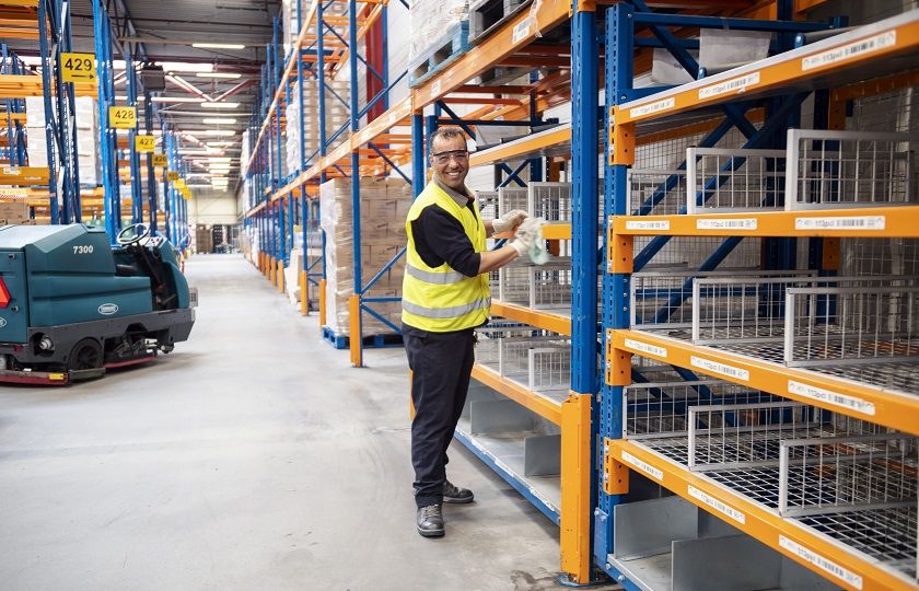 Een schoonmaakmedewerker van Gom Logistiek kijkt lachend in de camera en staat voor stellingen van een distributiecentrum