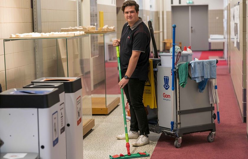 een medewerker van Gom Onderwijs kijkt in de camera en stofwist de vloer, met achter hem een werkwagen opgesteld