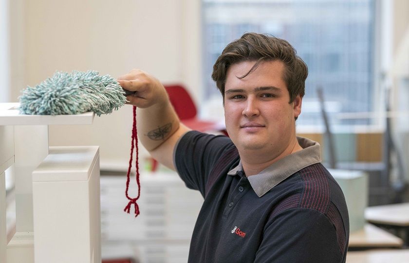 een medewerker van Gom Onderwijs kijkt in de camera en wist met een stofwisser de stof van een maquette