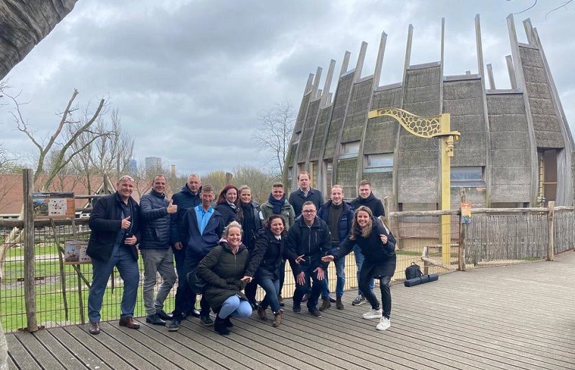 Een groep rayonleiders kijken je lachend aan en staan voor het verblijf van de giraffen in Blijdorp