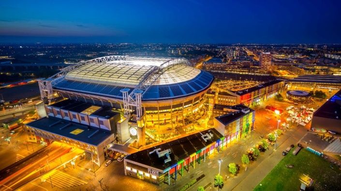 De kijk van bovenaf op de  Johan Cruijff Arena in Amsterdam in de avond waarbij zowel de arena als het treinstation is verlicht.